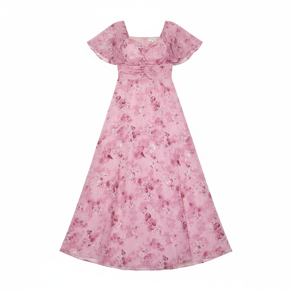 A woman models a pink floral maxi dress with short puff sleeves, a square neckline, and a gathered waist, standing in front of a white curtain backdrop with a wooden floor and a potted plant to the side.
