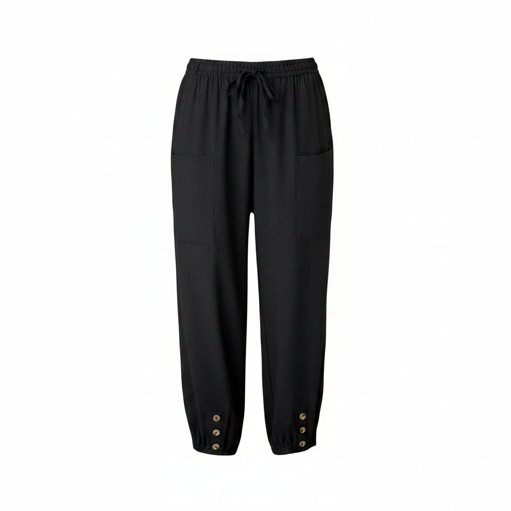 Black drawstring cropped pants with side pockets and button details at the cuffs, shown on a model wearing brown sandals in a bright indoor setting.