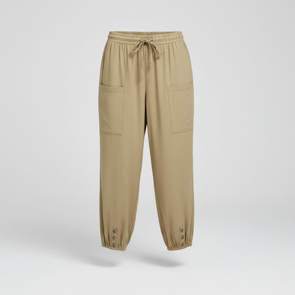 A person wearing loose beige drawstring pants with large front patch pockets and gathered cuffed hems, shown from the waist down against a light indoor wall and wood floor.