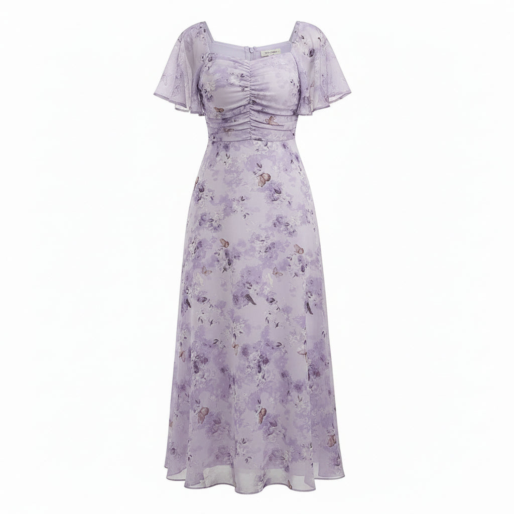 A woman models a lavender floral maxi dress with a square neckline, short puff sleeves, and a gathered ruched bodice. She is standing in a room with a white curtain background, a wooden floor, and a potted plant on the left.