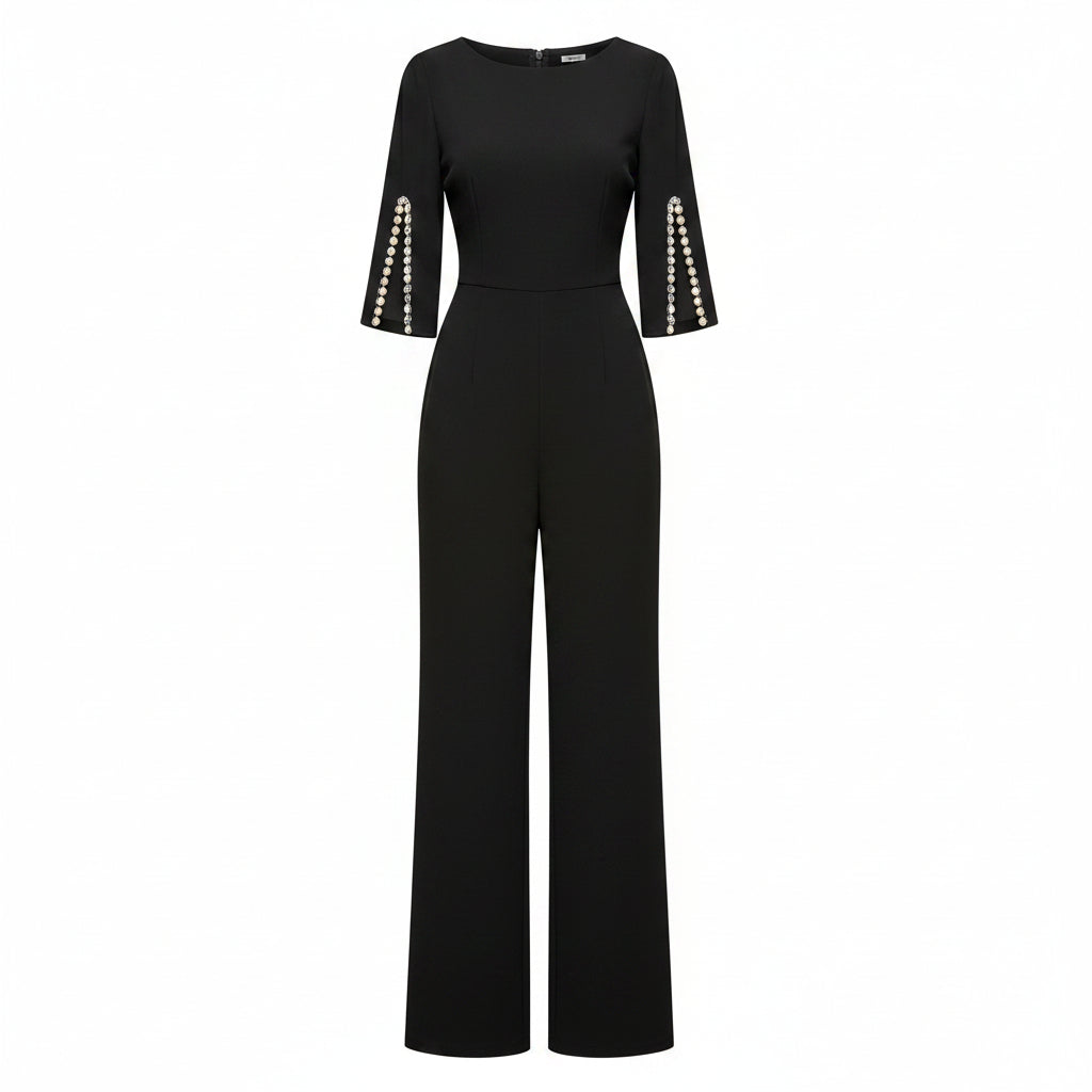 Woman wearing a black jumpsuit with pearl shoulder details against a plain wall.